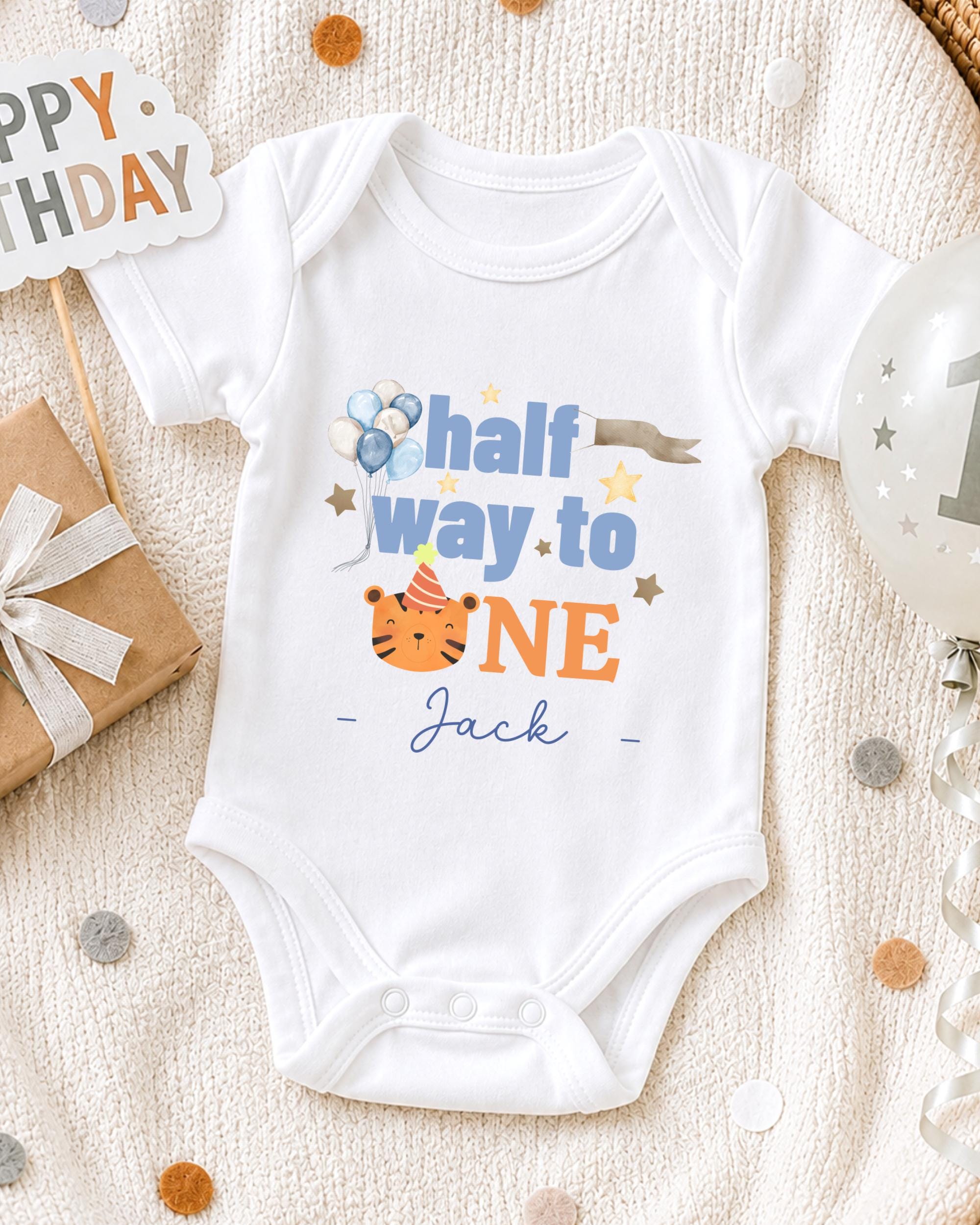 A white onesie with a personalized message, "Half way to one Jack", is displayed on a bed with a "Happy Birthday" sign and a birthday balloon.