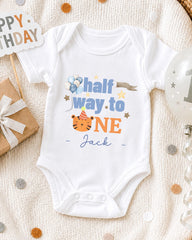 A white onesie with a personalized message, "Half way to one Jack", is displayed on a bed with a "Happy Birthday" sign and a birthday balloon.