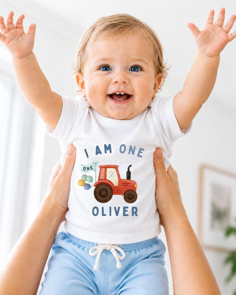 A smiling baby wearing a white shirt with a tractor graphic and the text "I am one Oliver" is being held up by someone's arms.