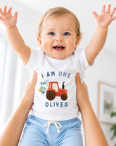 A smiling baby wearing a white shirt with a tractor graphic and the text "I am one Oliver" is being held up by someone's arms.