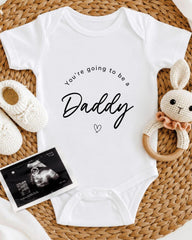 A white baby onesie with the text "You're going to be a Daddy" printed on it, along with a pair of white crocheted shoes and a stuffed bunny toy.