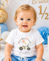 A smiling baby boy wearing a white t-shirt with a birthday-themed graphic and the text "Half way to one Hunter" is sitting in front of a birthday party backdrop.