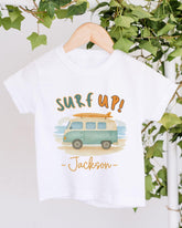 A white t-shirt with a graphic design of a van and the text "Surf up! Jackson" printed on it, hanging on a wooden hanger.