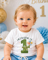 A smiling baby boy wearing a white t-shirt with the text "JACKSON IS 1" and a green number 1, sitting in front of a birthday party backdrop with balloons and confetti.