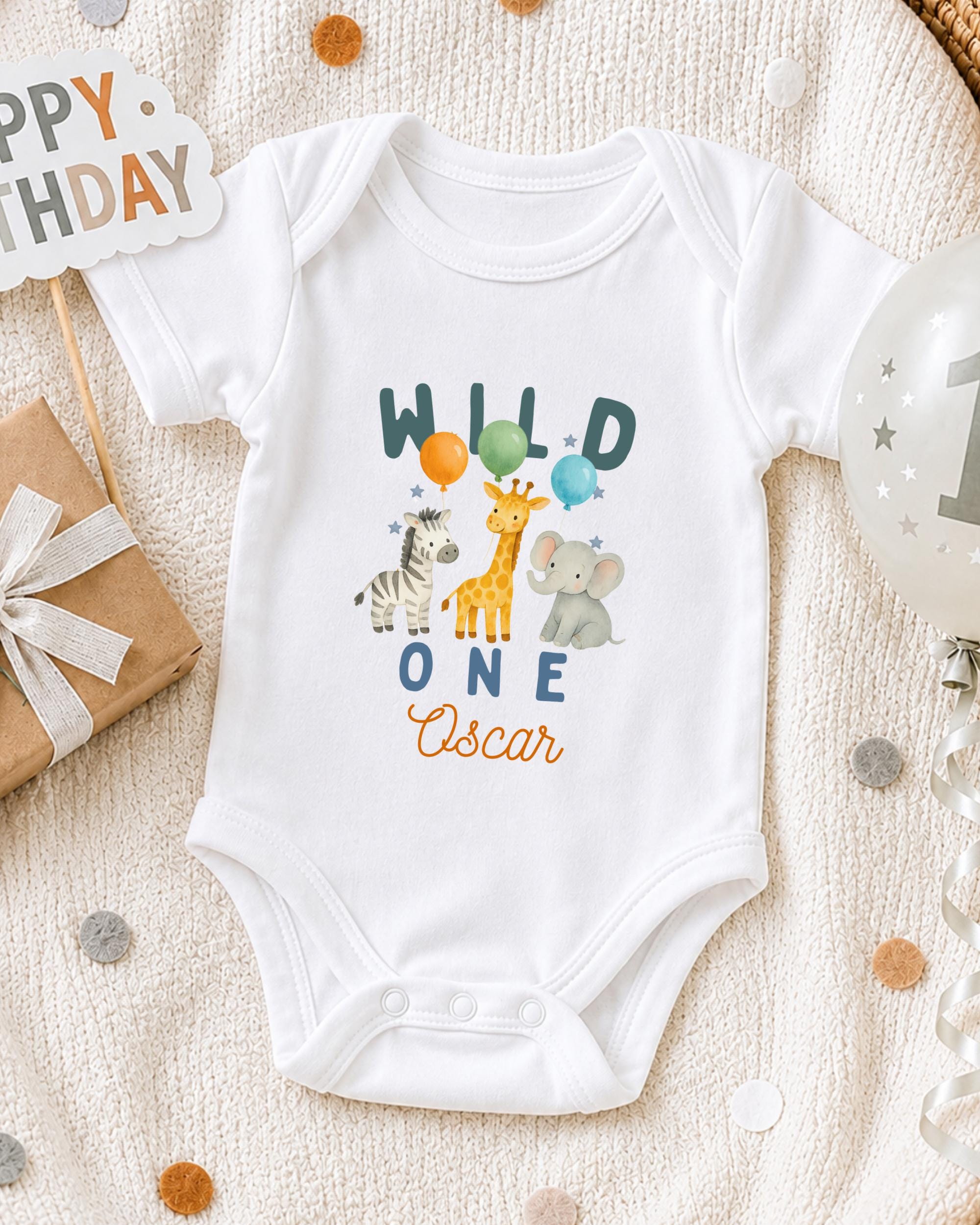A white onesie with a design of wild animals, including a giraffe, zebra, and elephant, along with the text "Wild One Oscar" printed on it. The onesie is placed on a white blanket, surrounded by various party decorations such as balloons and a "Happy Birthday" sign.