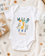A white onesie with a design of wild animals, including a giraffe, zebra, and elephant, along with the text "Wild One Oscar" printed on it. The onesie is placed on a white blanket, surrounded by various party decorations such as balloons and a "Happy Birthday" sign.
