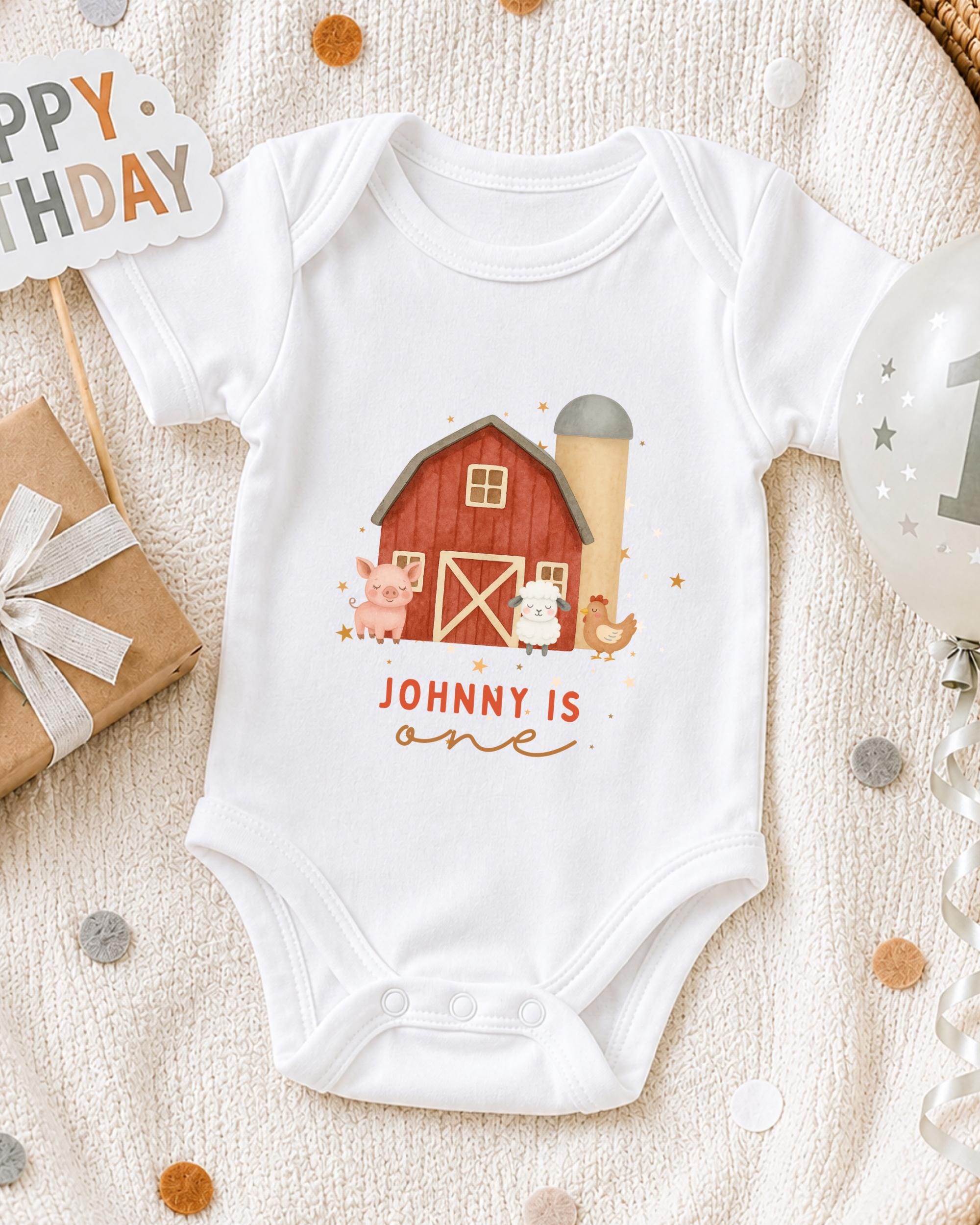 A white onesie with a farm scene and the text "JOHNNY IS ONE" printed on it, along with a red barn and other farm animals, is displayed on a bed with a "Happy Birthday" sign and a balloon.