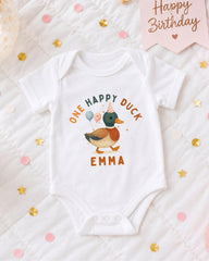 A white onesie with a cartoon duck and the text "One Happy Duck Emma" printed on it, surrounded by confetti and a birthday card.