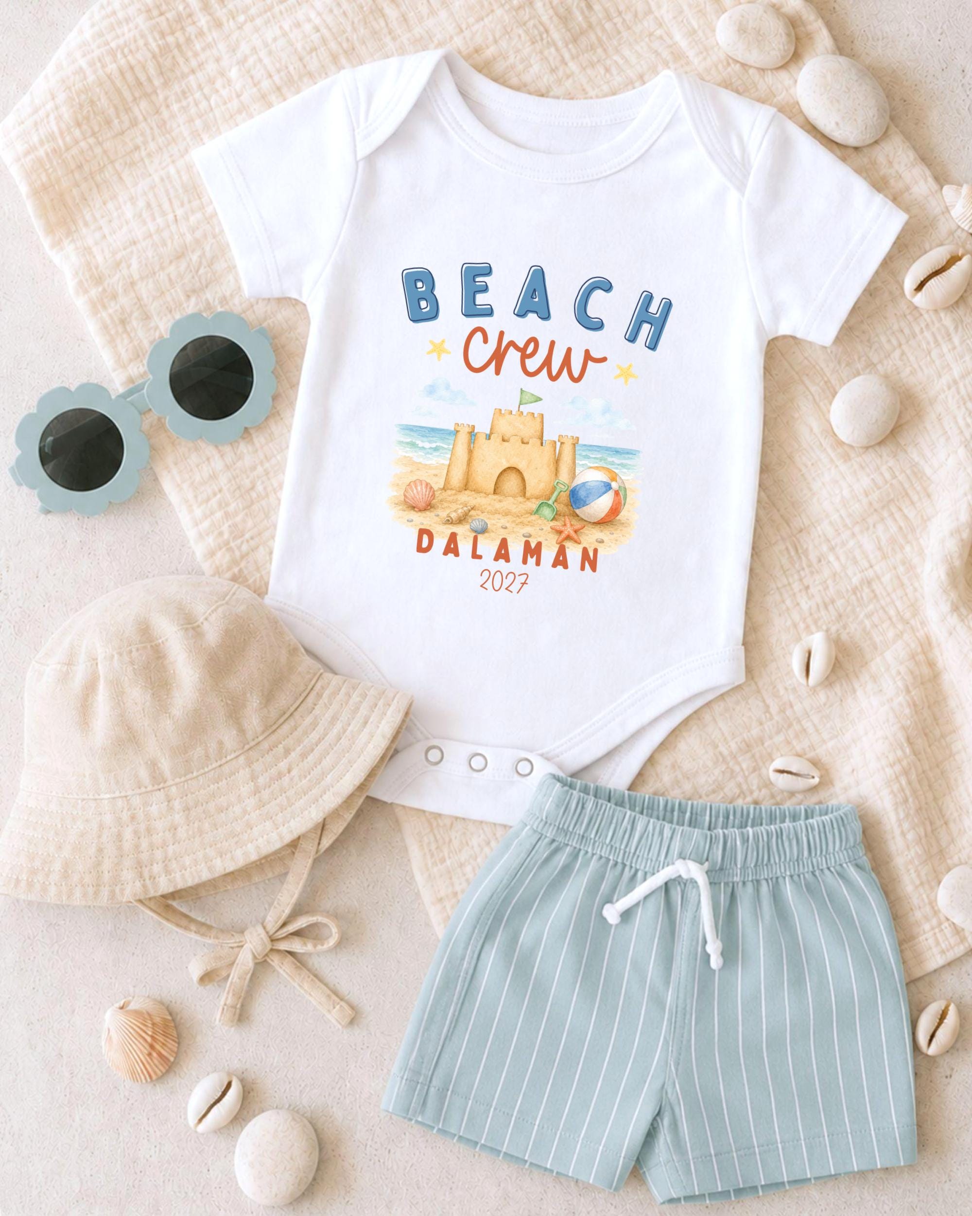A white baby onesie with a beach crew design, a beige bucket hat, a pair of blue shorts, and a pair of sunglasses are arranged on a beige surface.