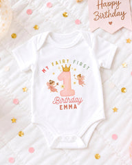 A white onesie with a birthday message and a fairy design, surrounded by confetti and a birthday card.