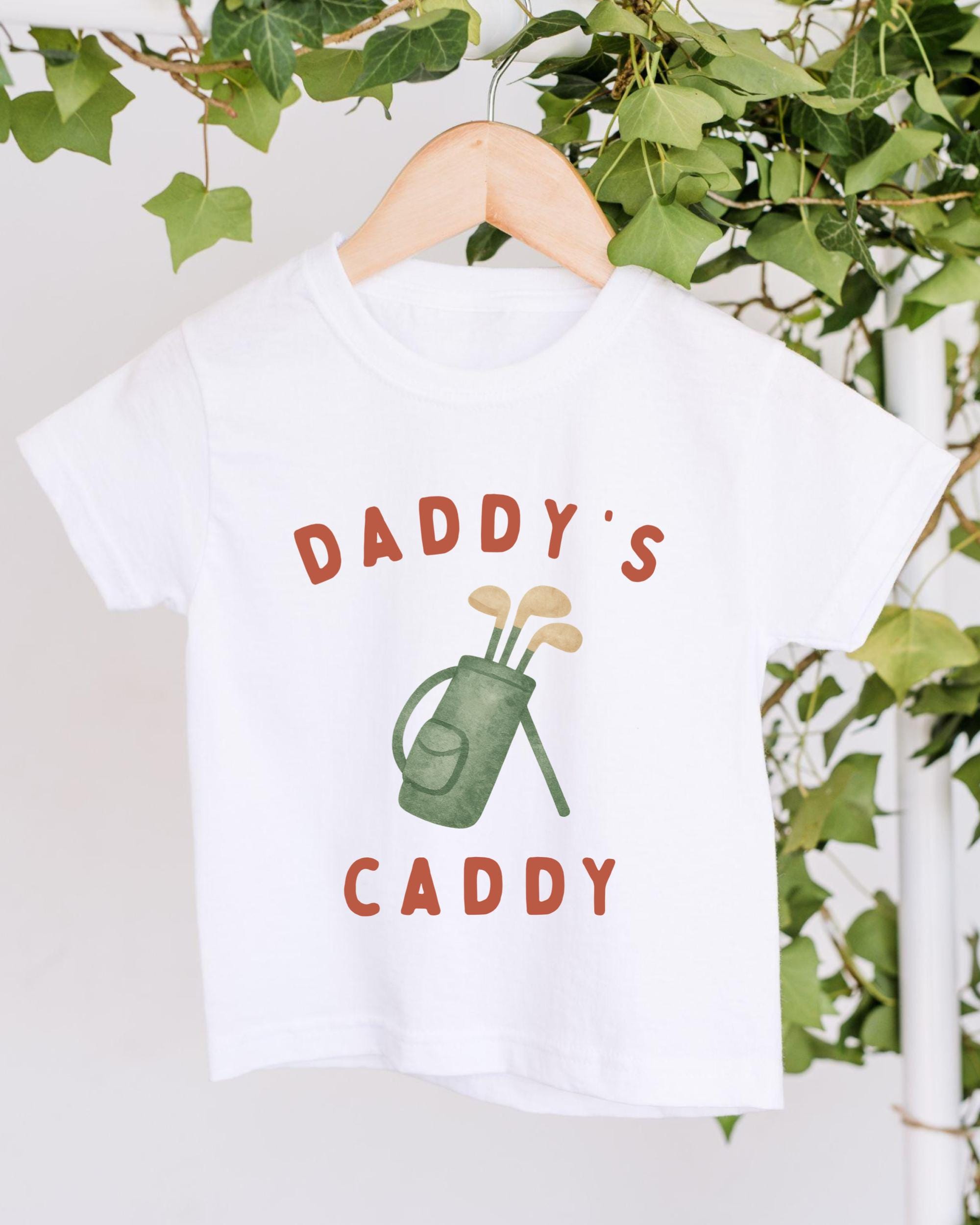 A white t-shirt with a graphic design of a golf bag and the text "DADDY'S CADDY" printed on it, hanging on a wooden hanger.