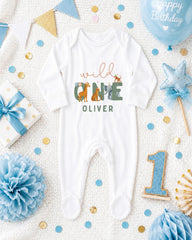 A white onesie with the text "wild one Oliver" printed on it, surrounded by various baby-themed items such as a blue balloon, a star, and confetti.
