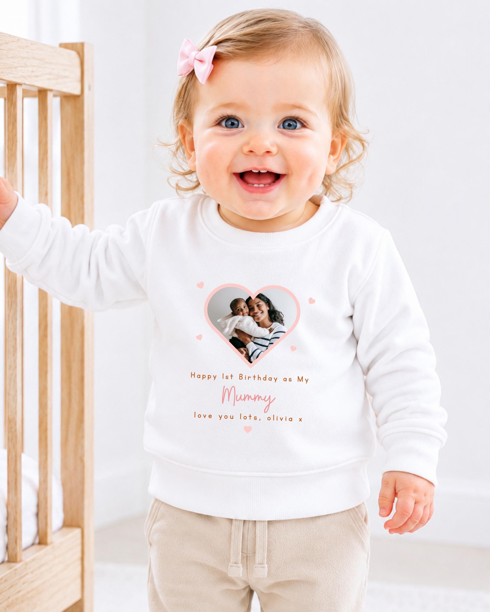 A smiling baby girl wearing a white sweatshirt with a heart-shaped photo and text on it.