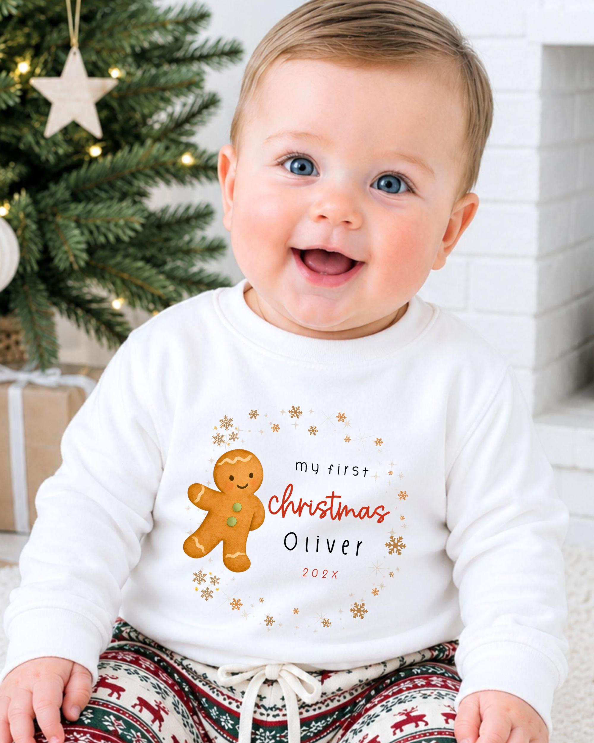 A smiling baby boy wearing a white sweatshirt with a gingerbread man and text that says "My first Christmas Oliver" and "2021".