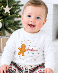 A smiling baby boy wearing a white sweatshirt with a gingerbread man and text that says "My first Christmas Oliver" and "2021".