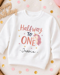 A white sweatshirt with a personalized text and a unicorn graphic printed on it, surrounded by confetti.