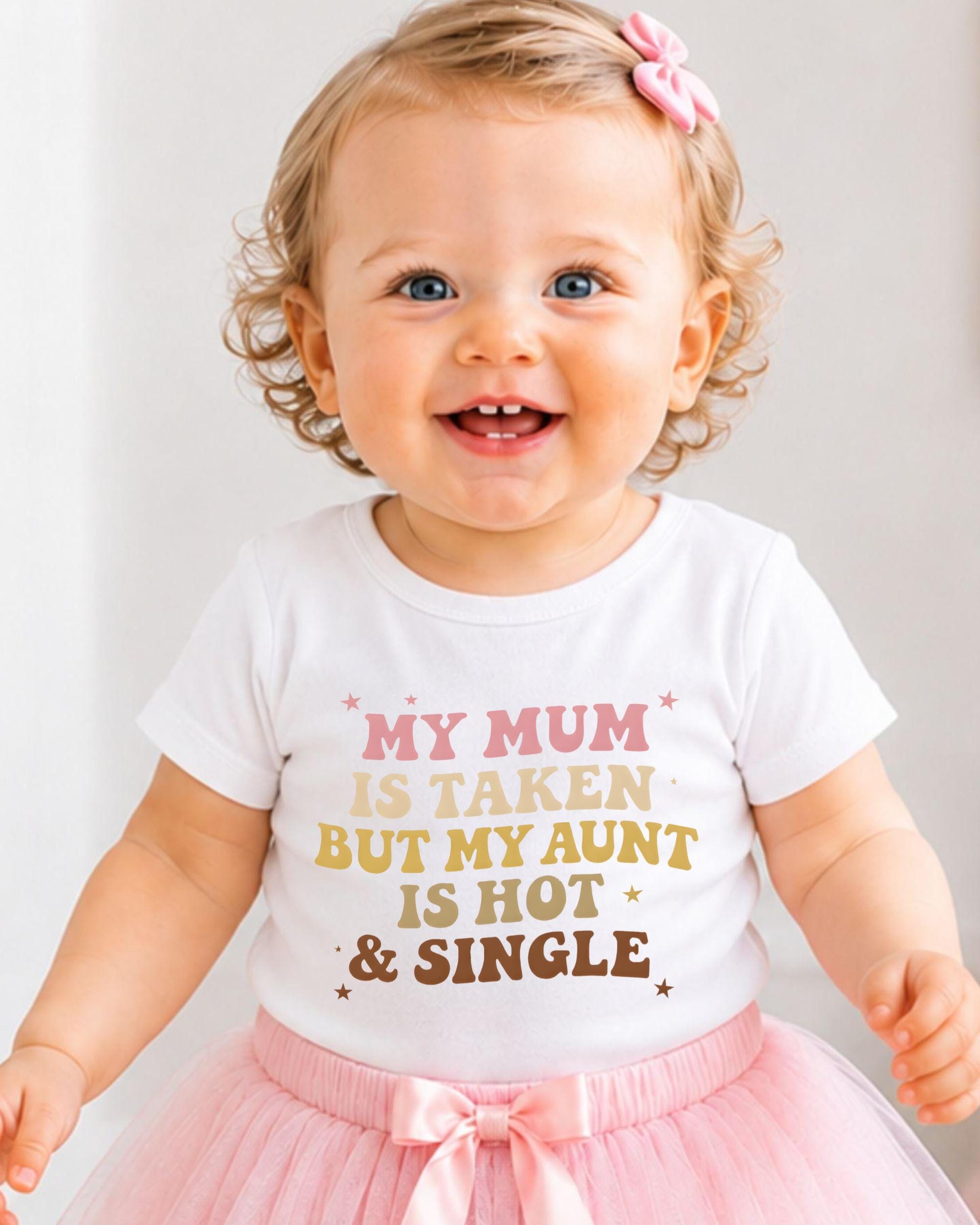 A smiling baby girl wearing a white t-shirt with text and a pink bow in her hair.