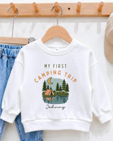 A white sweatshirt with a graphic design of a camping scene and the text "My first camping trip" printed on it, hanging on a wooden hanger.