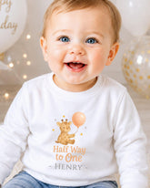 A smiling baby boy wearing a white shirt with a giraffe and balloon design, sitting in front of birthday balloons.