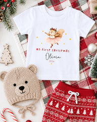 A white t-shirt with a cartoon image of a squirrel and the text "My first Christmas Olivia 2020" printed on it, along with a red and white patterned pair of shorts and a beige knit hat. The t-shirt is surrounded by Christmas decorations, including a small Christmas tree, ornaments, and a red and white plaid blanket.
