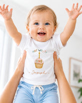 A smiling baby wearing a white t-shirt with a cupcake and the text "Half Way to One Joseph" is being held up by someone's arms.