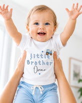 A smiling baby wearing a white shirt with the text "Little Brother Oscar" is being held up by someone's arms.