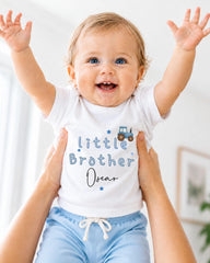 A smiling baby wearing a white shirt with the text "Little Brother Oscar" is being held up by someone's arms.