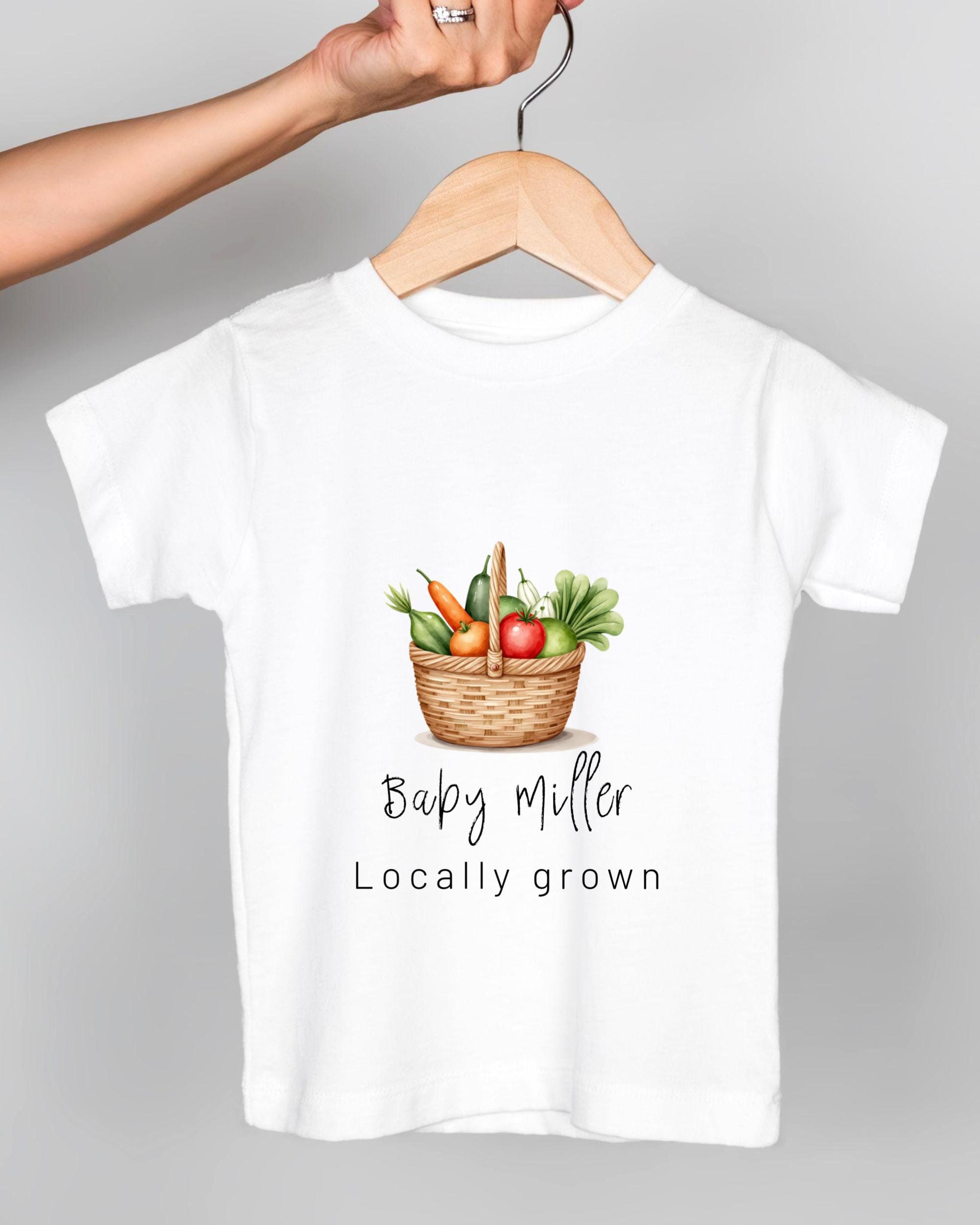 A white t-shirt with a graphic design of a basket filled with fresh vegetables, including carrots, tomatoes, and lettuce. The text below the image says "Baby Miller Locally grown".