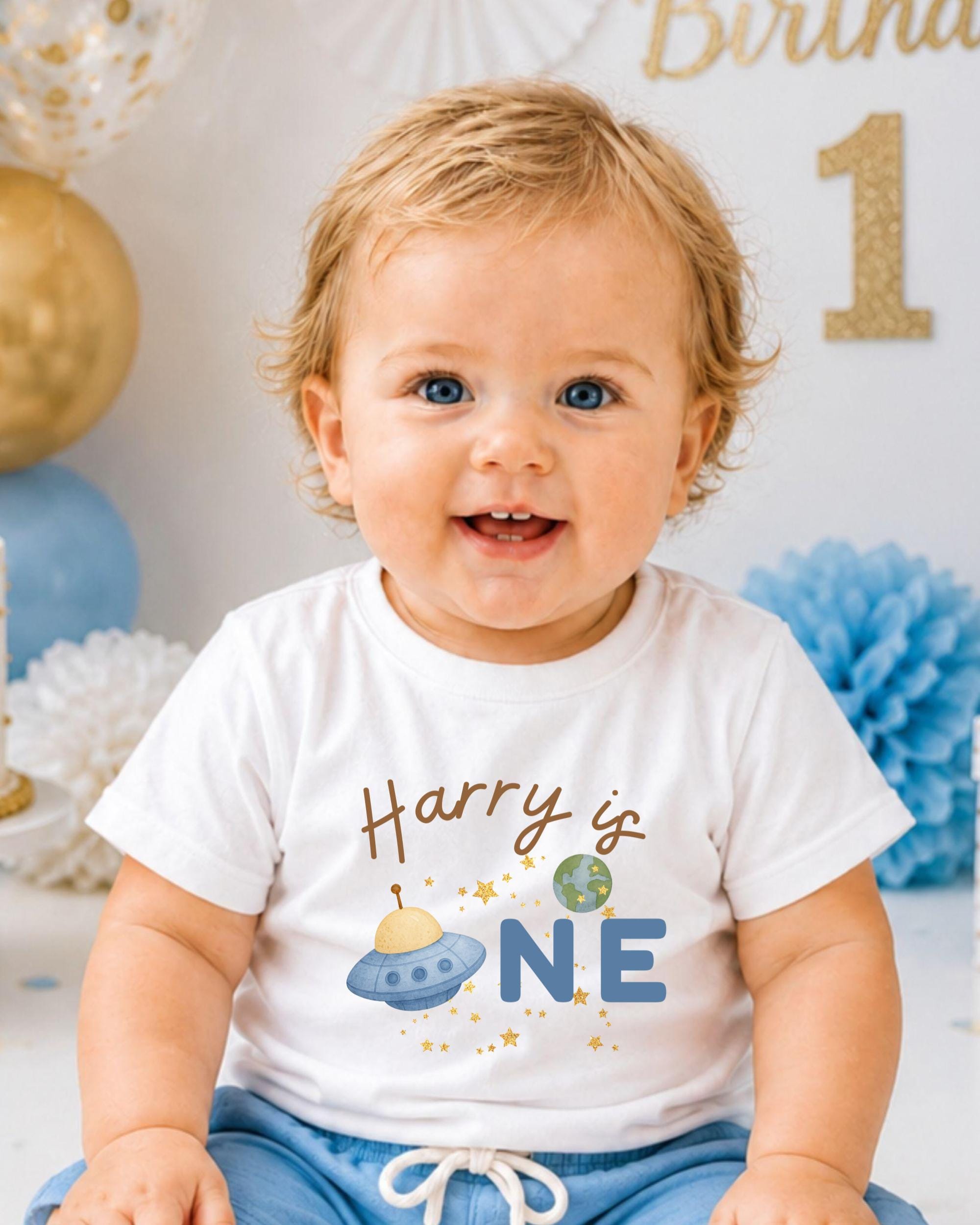 A smiling baby boy wearing a white t-shirt with the text "Harry is one" and an illustration of a spaceship.