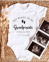 A white baby onesie with the text "GET READY TO BE Grandparents In January 2028" printed on it, along with a pair of black and white baby footprints. The onesie is placed next to a set of three black and white baby ultrasound images.