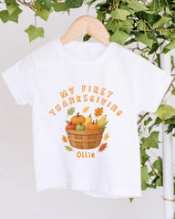 A white t-shirt with a graphic design featuring a basket filled with autumn fruits and vegetables, along with the text "My first Thanksgiving Ollie".
