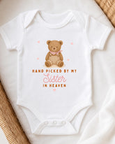 A white onesie with a teddy bear design and the text "Hand picked by my Sister in Heaven" printed on it.
