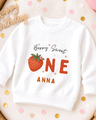 A white sweatshirt with a strawberry design and text printed on it, placed on a surface with confetti.