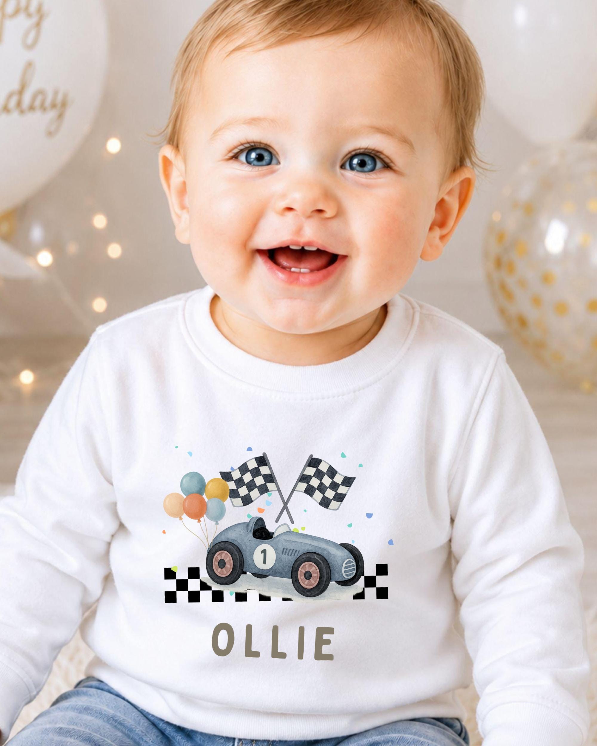 A smiling baby boy wearing a white shirt with a race car and checkered flags printed on it.