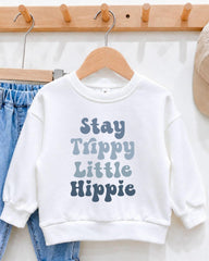 A white sweatshirt with the text "Stay Trippy Little Hippie" printed on it, hanging on a wooden hanger.