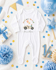 A white onesie with a car and balloons, surrounded by birthday decorations such as a blue balloon, blue and gold star, and blue and gold confetti.