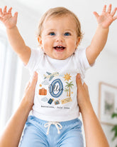 A smiling baby wearing a white shirt with a graphic design and text, being held up by someone's arms.