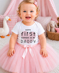 A smiling baby girl wearing a white t-shirt with text and a pink tutu, sitting on a pink carpet with stuffed animals in the background.