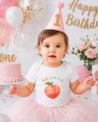 A young girl wearing a white t-shirt with a peach graphic and a pink birthday hat sits in front of a pink and white birthday cake, surrounded by balloons and flowers.