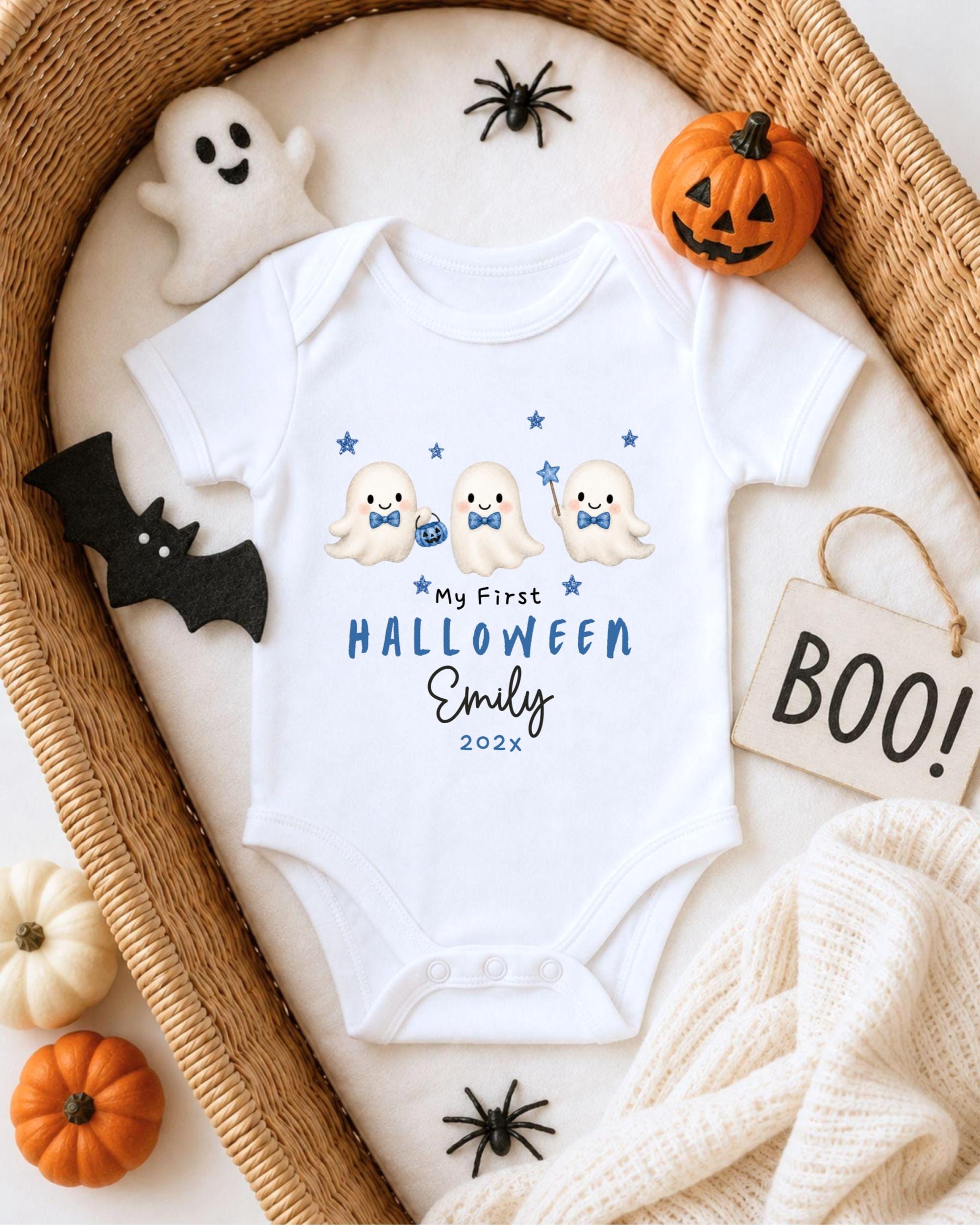 A white onesie with a Halloween theme, featuring three cute ghost characters, is displayed in a wicker basket. The onesie has a personalized text that reads "My First Halloween Emily 2023". The basket is surrounded by Halloween decorations, including a carved pumpkin, a ghost, and a bat.