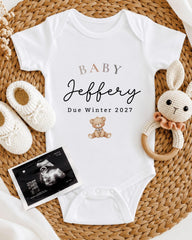 A baby announcement t-shirt with the name "Baby Jeffery" and the date "Due Winter 2027" printed on it, along with a teddy bear and a pair of crocheted baby booties. There is also an Ultrasound image of a baby's belly.