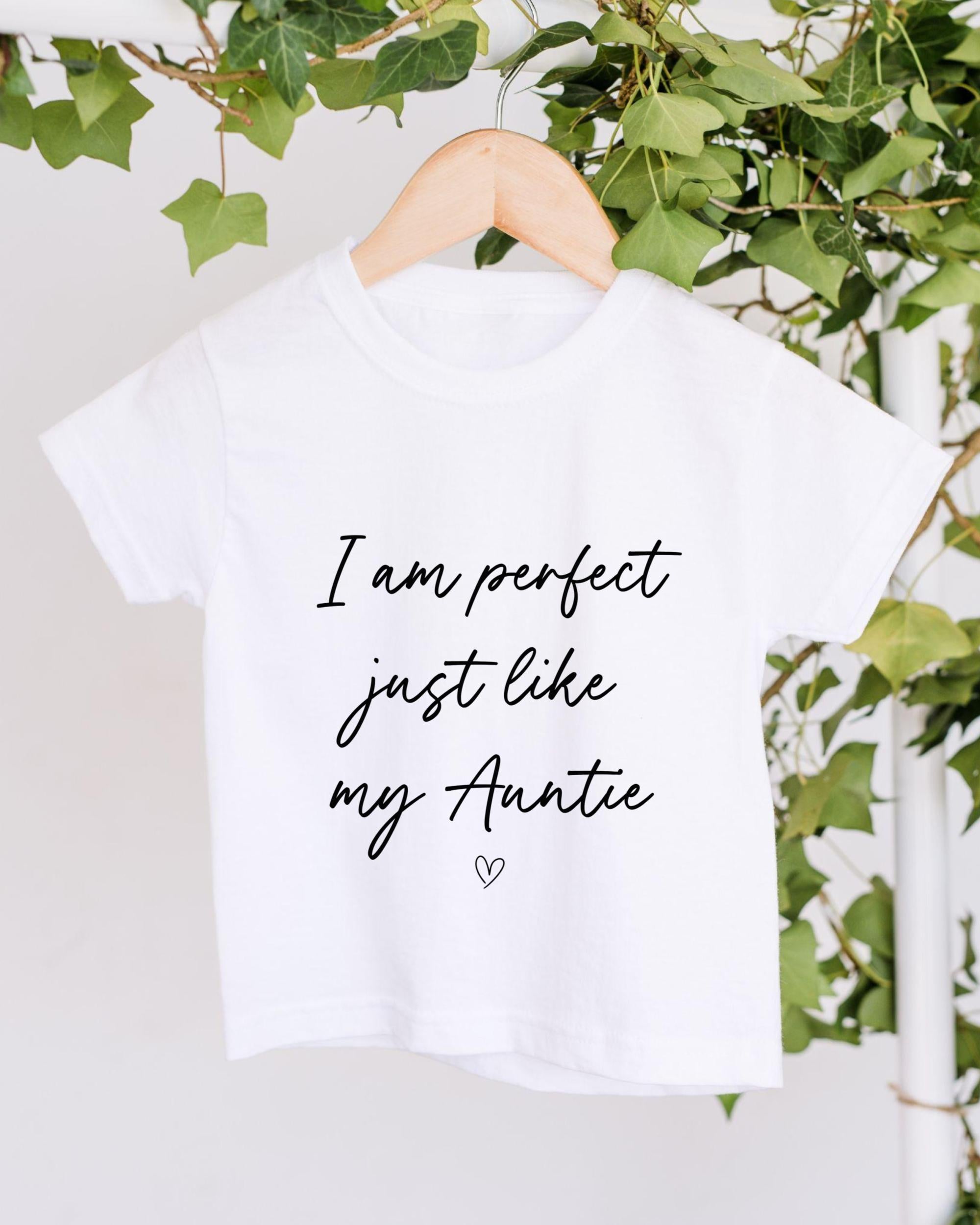 A white t-shirt with black text and a heart symbol hangs on a wooden hanger, with green ivy leaves and a white wall in the background.