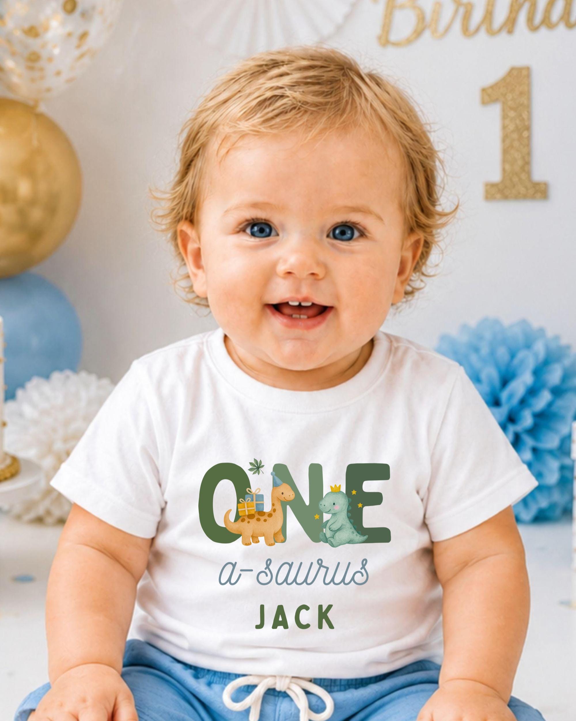 A smiling baby boy wearing a white t-shirt with dinosaur graphics and the text "QUE a-saurus JACK" is sitting in front of a birthday party backdrop.