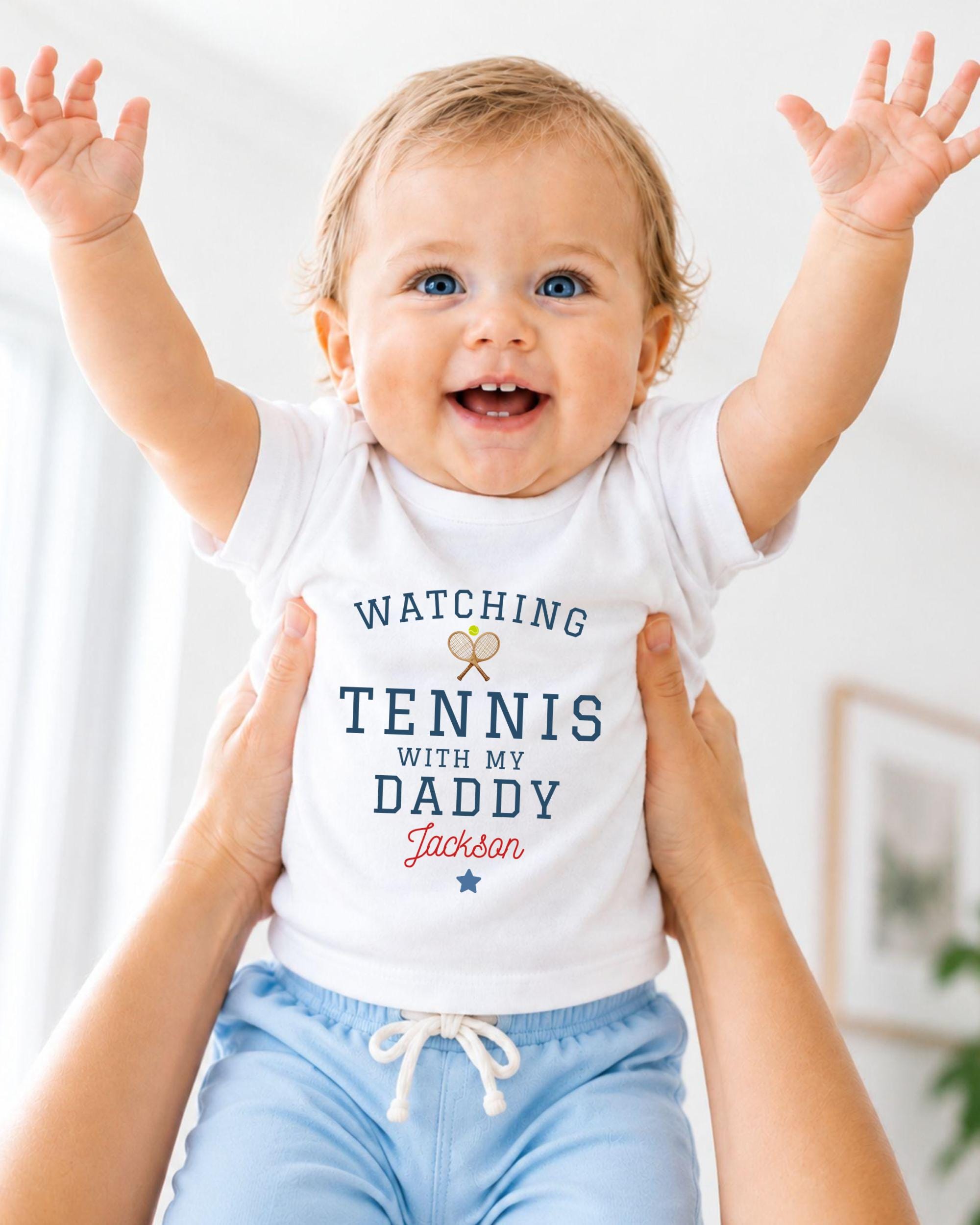 A smiling baby wearing a white shirt with the text "WATCHING TENNIS WITH MY DADDY JACKSON" is being held up by someone's arms.