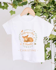 A white t-shirt with a graphic design featuring a baby deer and the text "Halfway to One 6 Months Christine" hanging on a wooden hanger.
