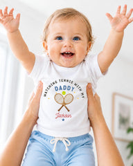 A smiling baby wearing a white shirt with a tennis racket and the text "Watching Tennis with my Daddy Jackson" is being held up by someone's arms.
