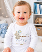 A smiling baby wearing a white onesie with a birthday message printed on it, sitting in a room with a teddy bear and bookshelf in the background.