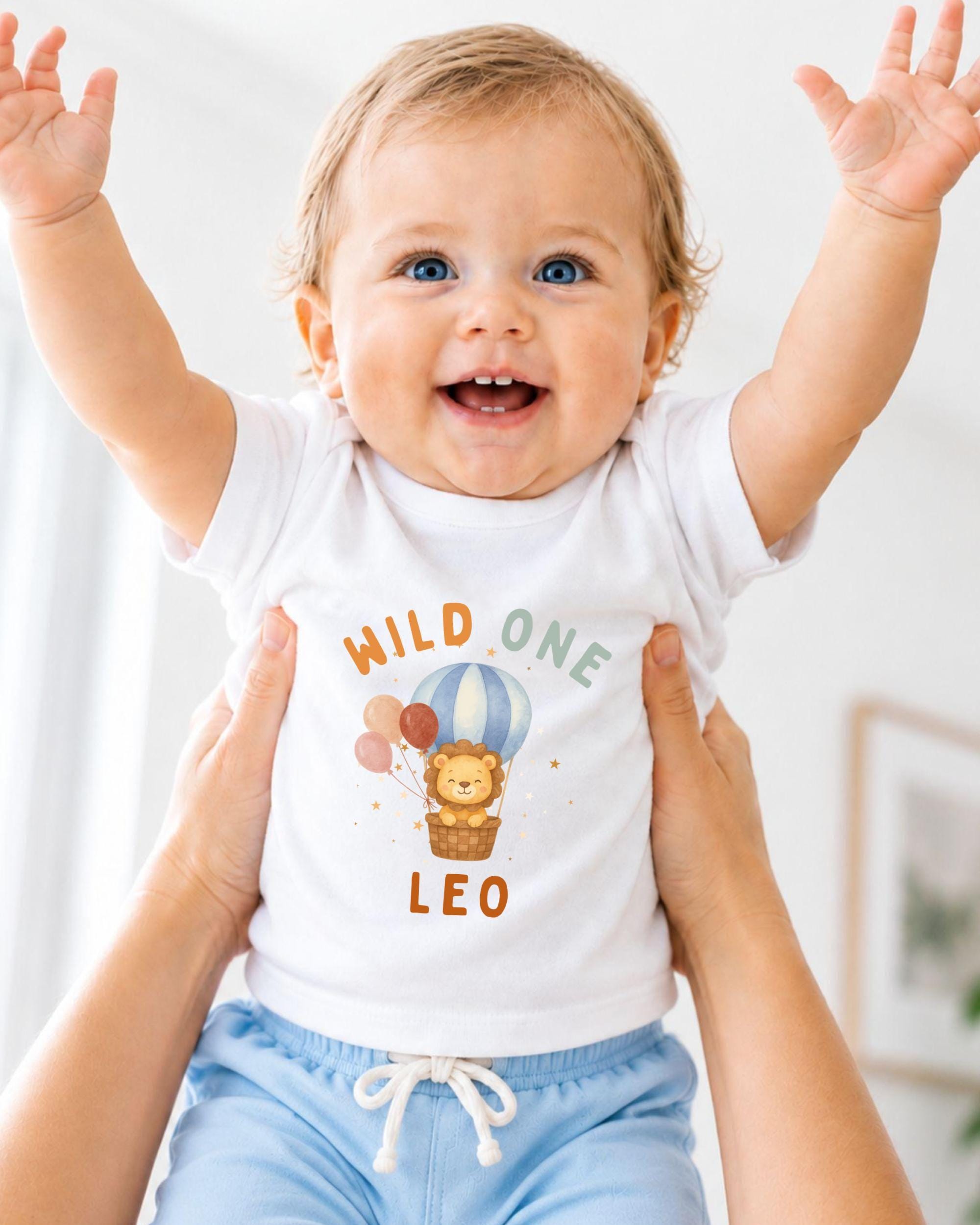 A smiling baby wearing a white t-shirt with a cartoon lion and balloons, being held up by someone's arms.