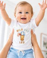 A smiling baby wearing a white t-shirt with a cartoon lion and balloons, being held up by someone's arms.