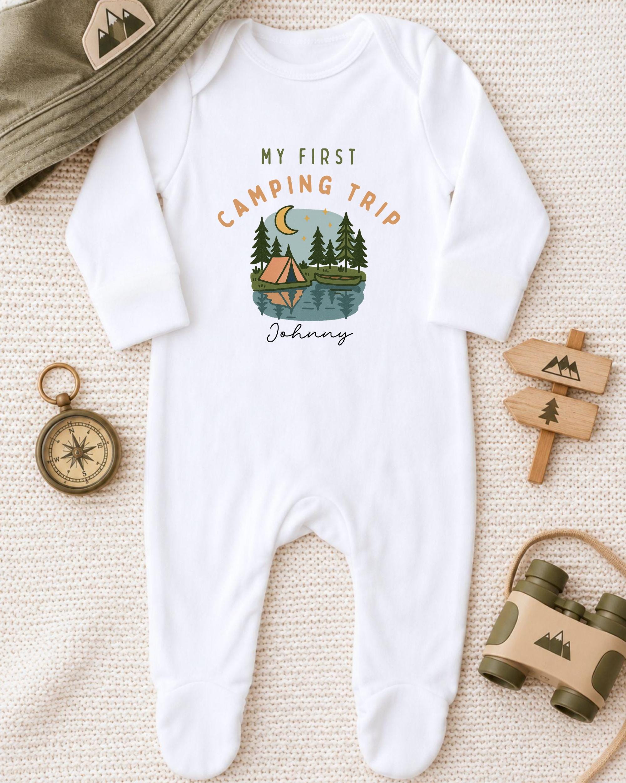 A white onesie with a camping-themed design, a compass, a hat, and a pair of binoculars, all arranged on a beige surface.